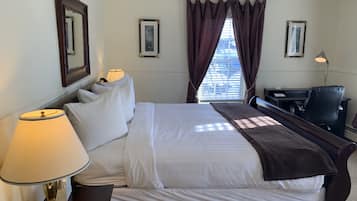 A neatly made bed with white linens and a brown throw, a desk with a lamp, and a chair in a hotel room with a window and curtains.