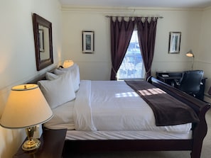 A neatly made bed with white linens and a brown throw, a desk with a lamp, and a chair in a hotel room with a window and curtains.