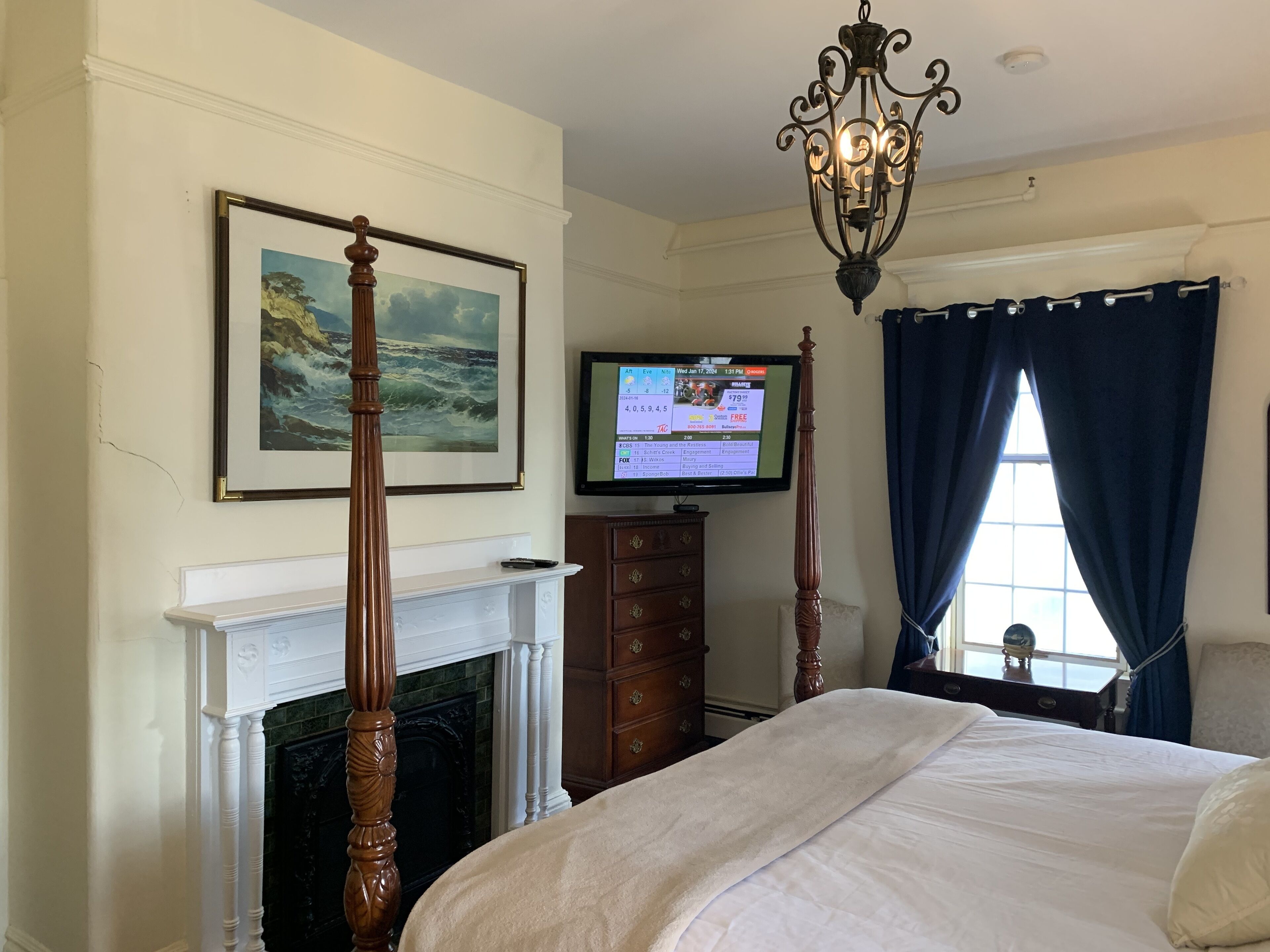 A bedroom with a four-poster bed, a fireplace, a television, and a window with curtains.