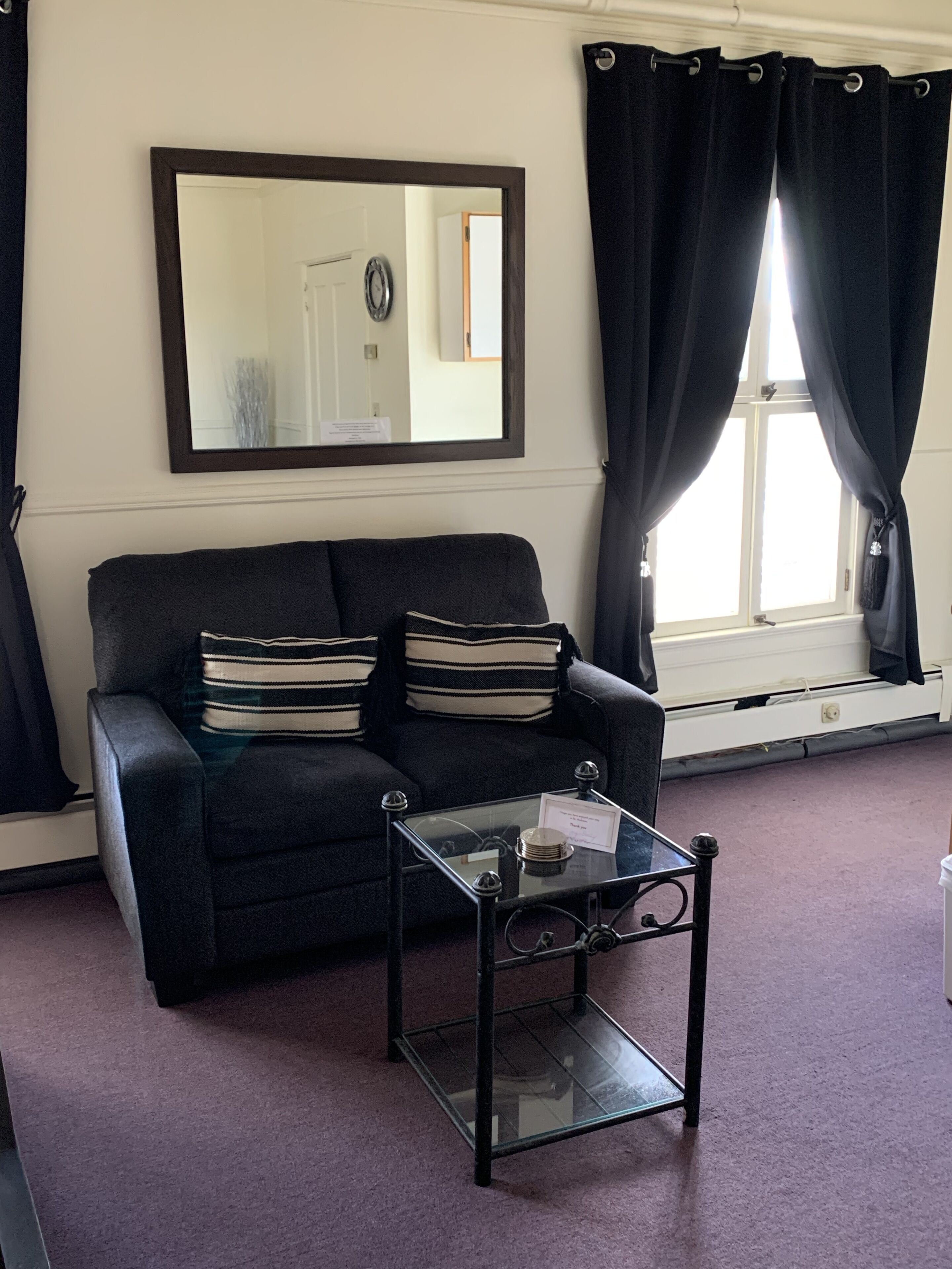 A living room with a dark sofa, striped cushions, a glass coffee table, a large mirror, and a window with curtains.