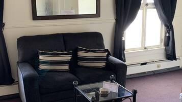 A living room with a dark sofa, striped cushions, a glass coffee table, a large mirror, and a window with curtains.