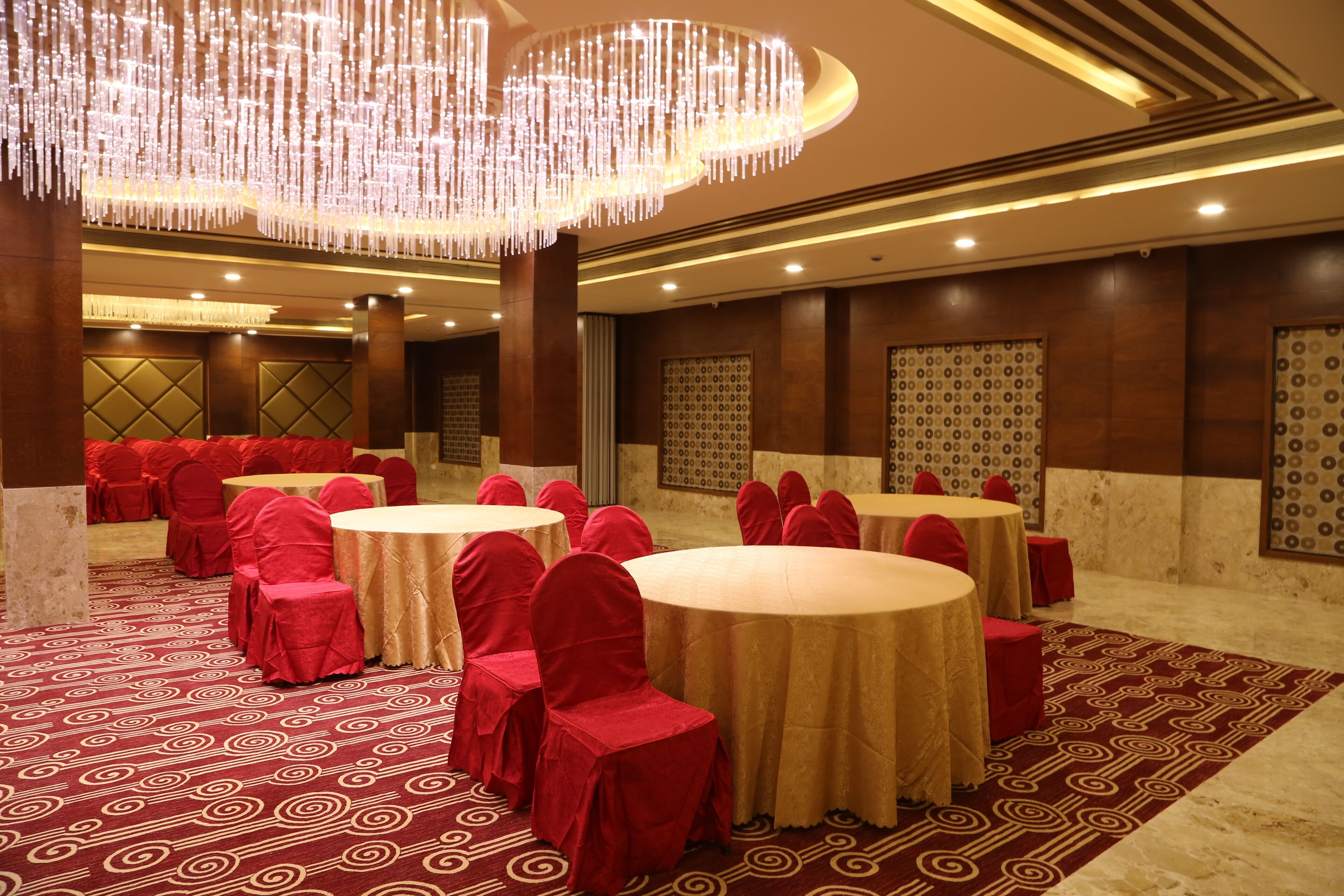 Une salle de banquet avec des tables rondes, des chaises recouvertes de tissu rouge et un tapis à motifs.