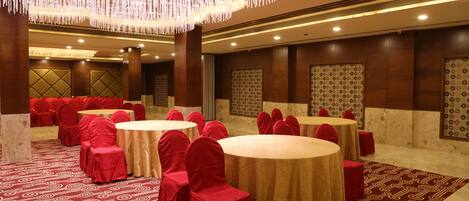 Une salle de banquet avec des tables rondes, des chaises recouvertes de tissu rouge et un tapis à motifs.