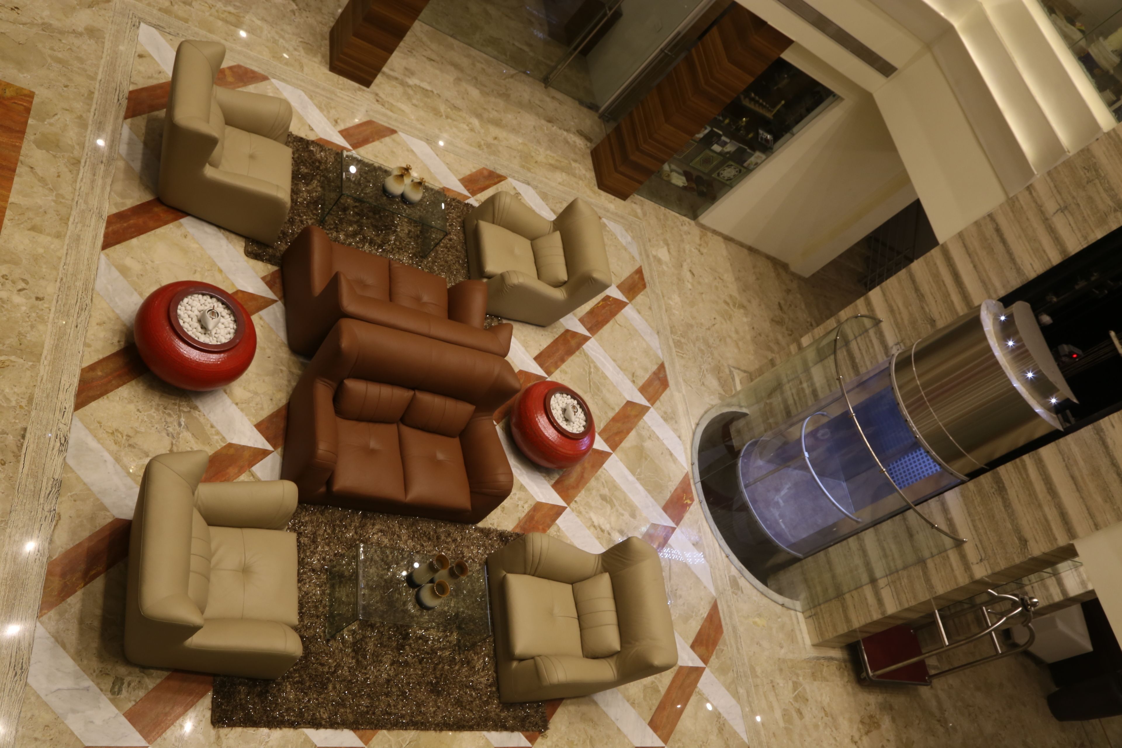 A lobby with a central seating area featuring a brown sofa and beige armchairs, a patterned floor, and a glass elevator.