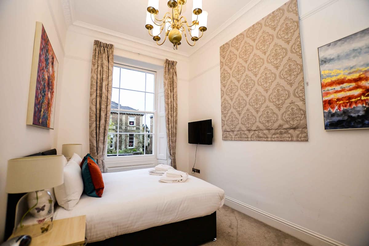 A hotel room with a large bed, a chandelier, a television, and a window with curtains.