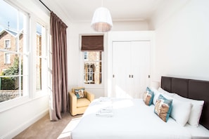 Une chambre avec un grand lit, une chaise, une fenêtre avec des rideaux et une vue sur les bâtiments à l’extérieur.