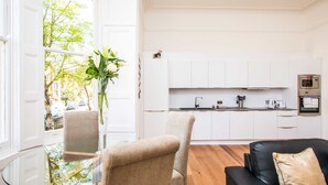 Una sala de estar moderna con una mesa de centro de vidrio, sillones color beige y un sofá negro con un cojín color beige. Se ve una cocina blanca con gabinetes blancos y electrodomésticos de acero inoxidable al fondo.