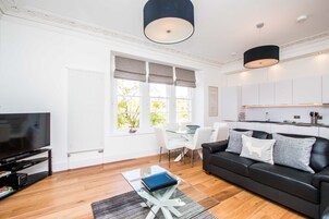 Una sala de estar moderna con televisor de pantalla plana, una mesa de centro de vidrio, un sofá de cuero negro y una zona de comedor con sillas blancas.
