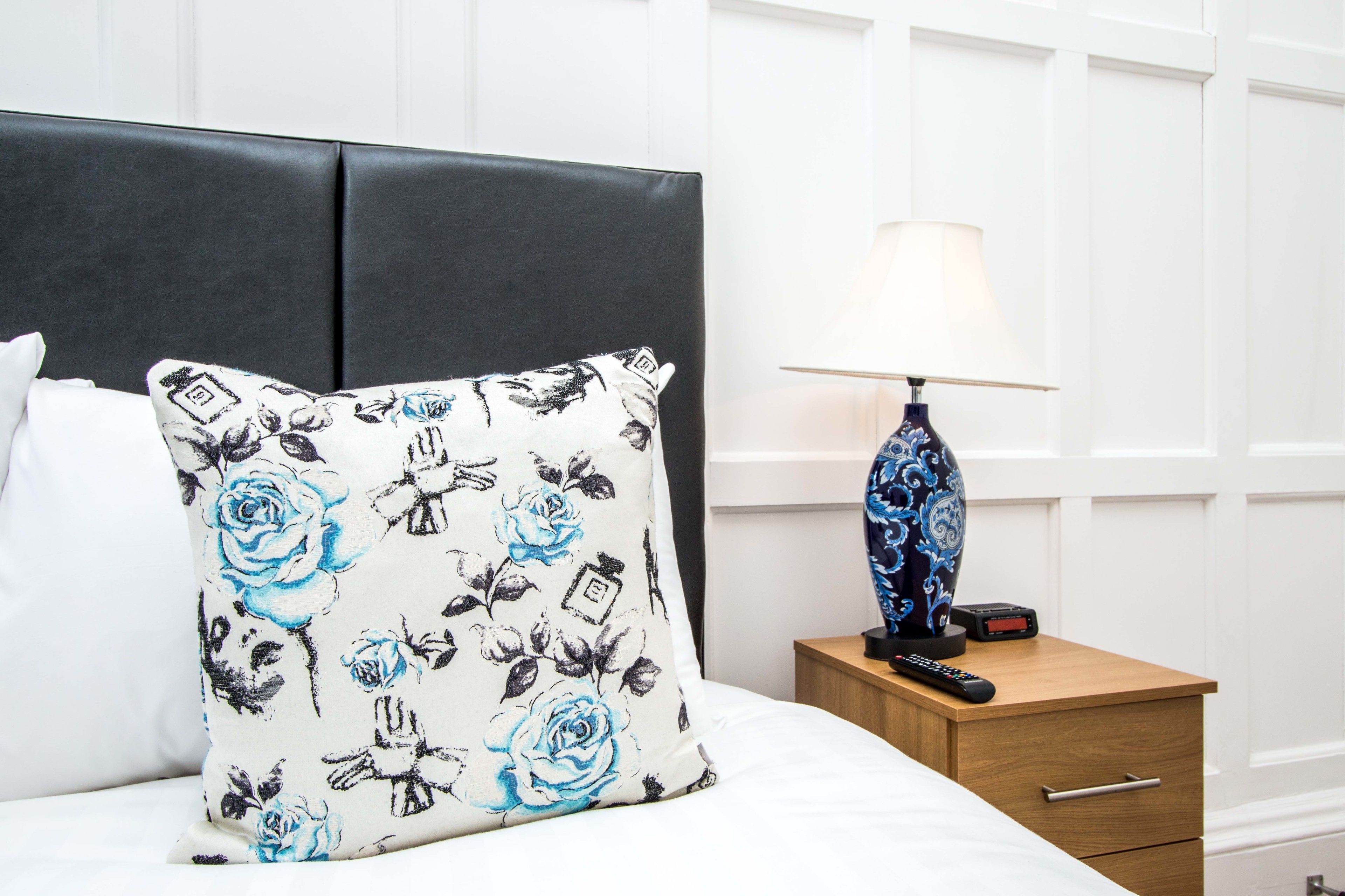 Una cama con un cojín floral, una lámpara de noche y una mesita de madera con un control remoto.