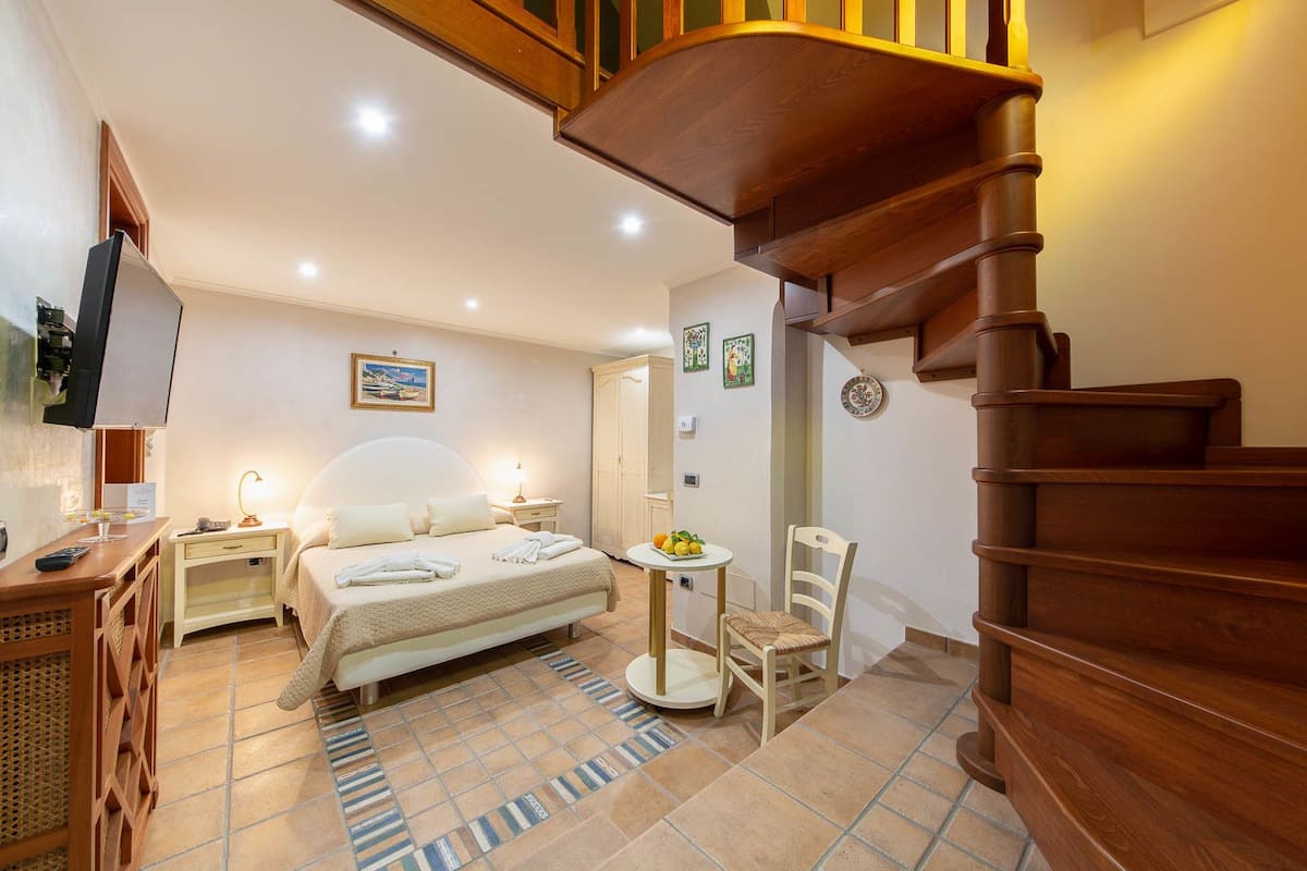 Une chambre d’hôtel avec un lit, une table de chevet, une lampe, une chaise, une petite table avec des fruits et un escalier en bois.