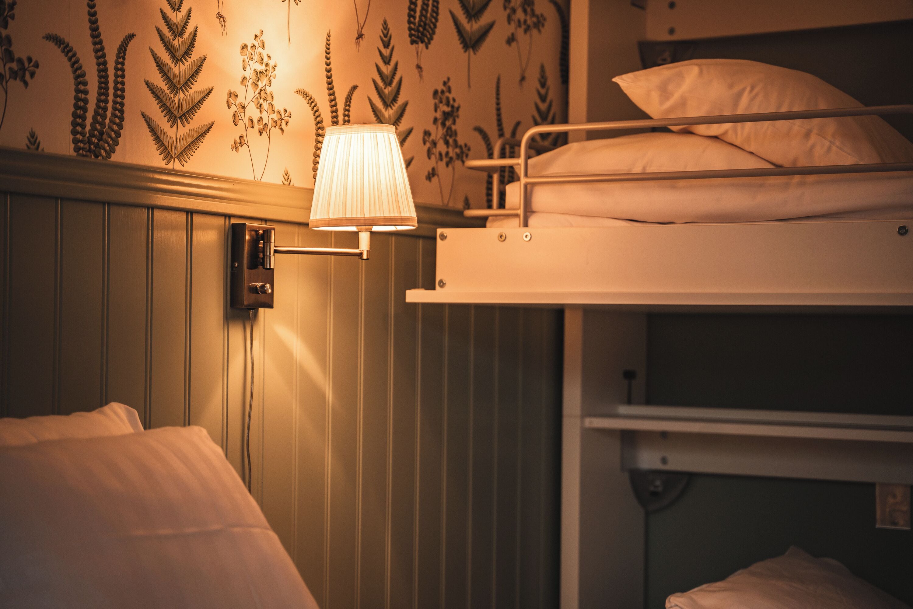 A bunk bed with a wall-mounted lamp, a power outlet, and a floral wallpaper design.