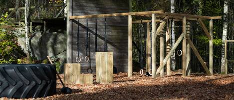 A wooden structure with a climbing wall and gymnastic rings in a forested area.