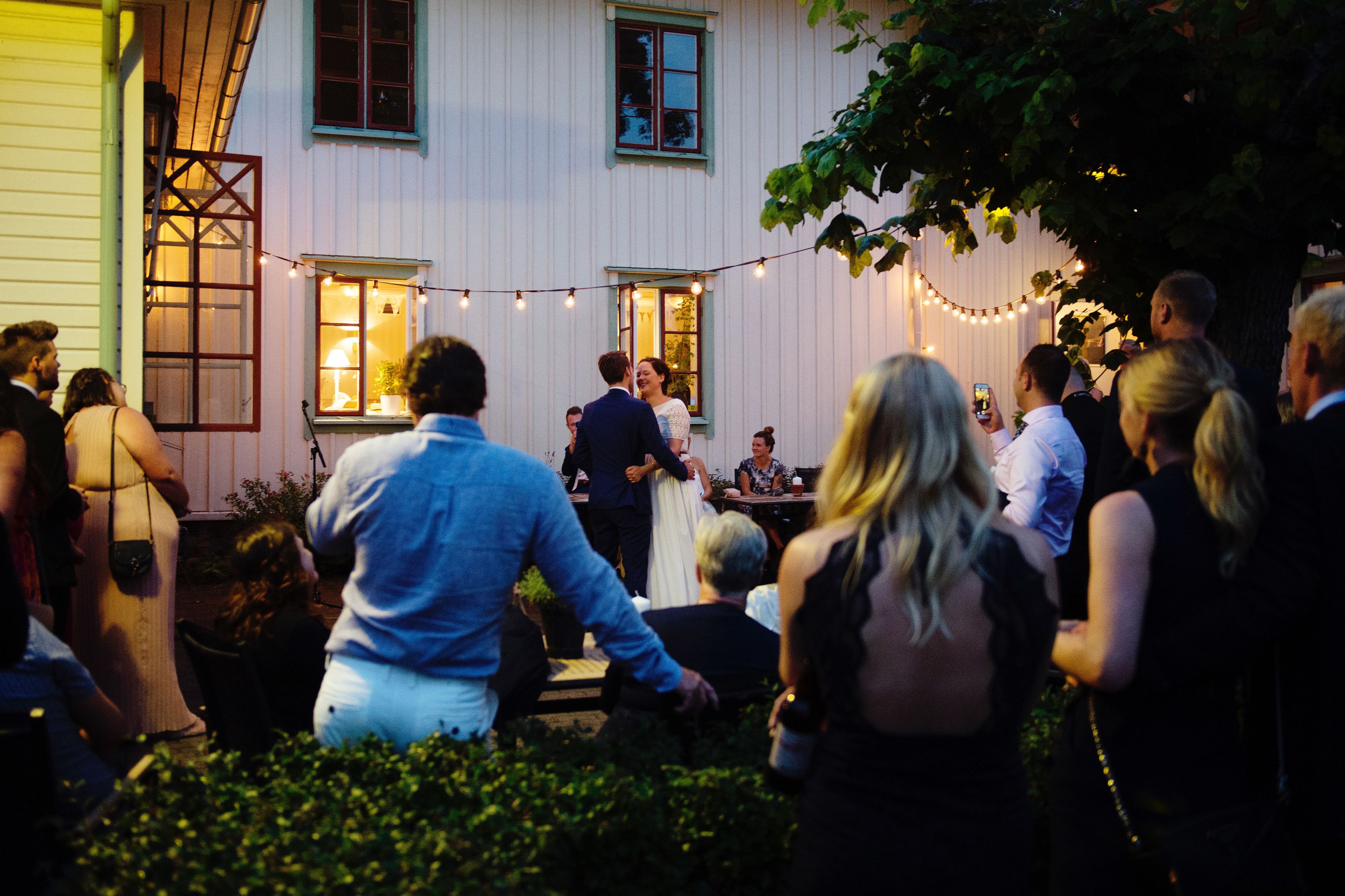 A group of people gathered outside a building with string lights, a window with a lamp, and a door with a lattice design.
