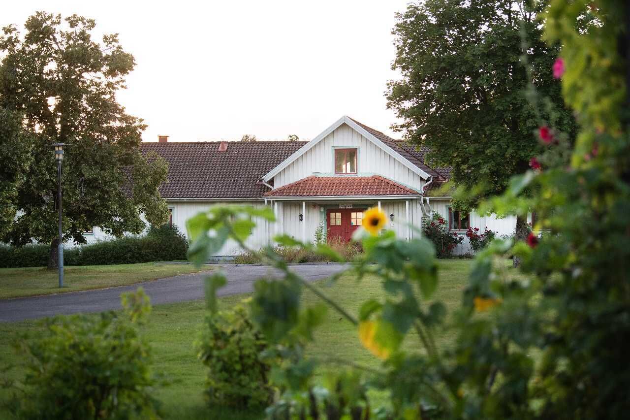A white house with a red door and a tiled roof, surrounded by greenery and trees.