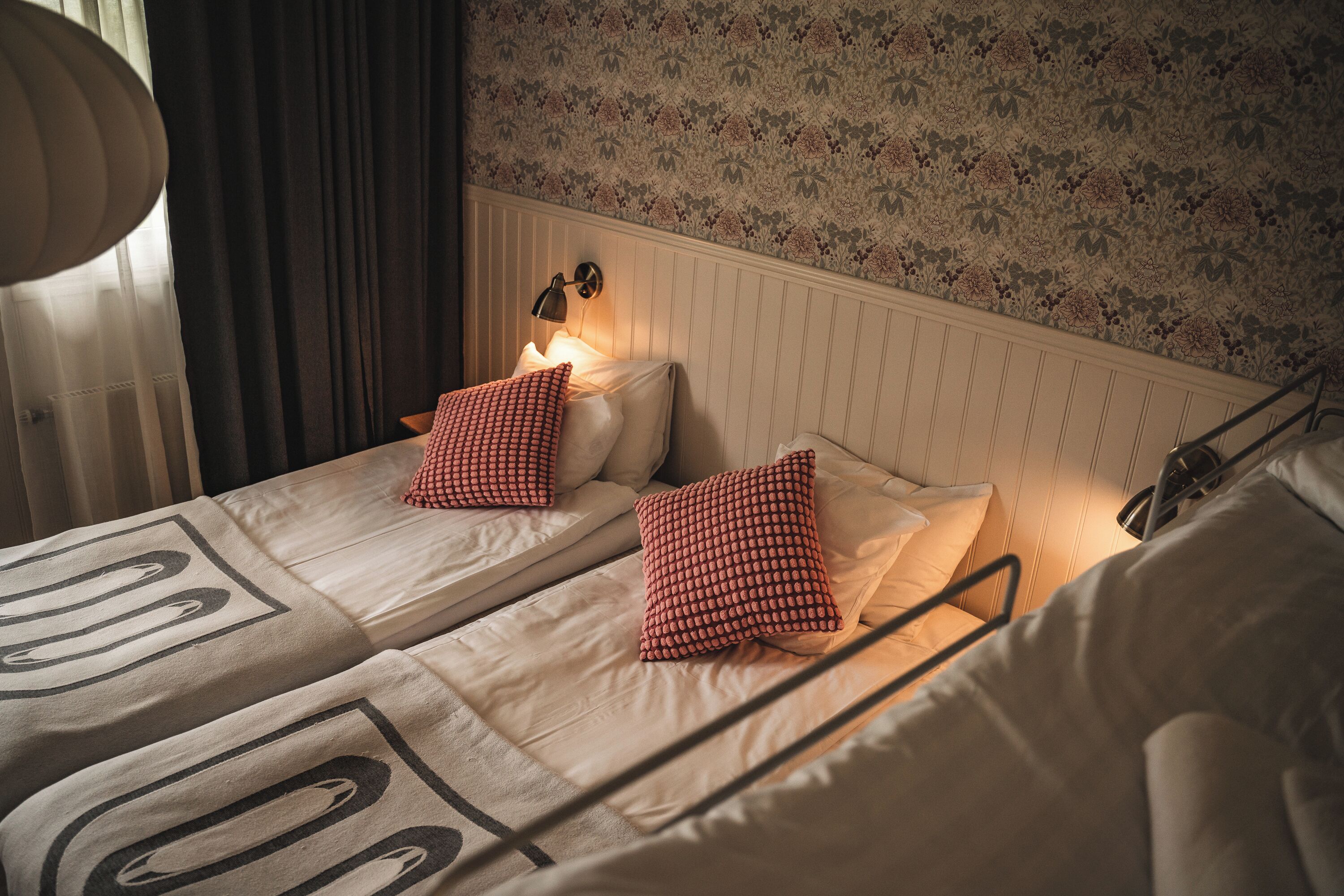 A hotel room with two beds, a nightstand with a lamp, and a floral-patterned wall.