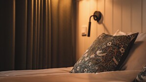 A neatly made bed with a patterned pillow, a wall-mounted lamp, and a window with curtains. - Två Skyttlar (Orby)