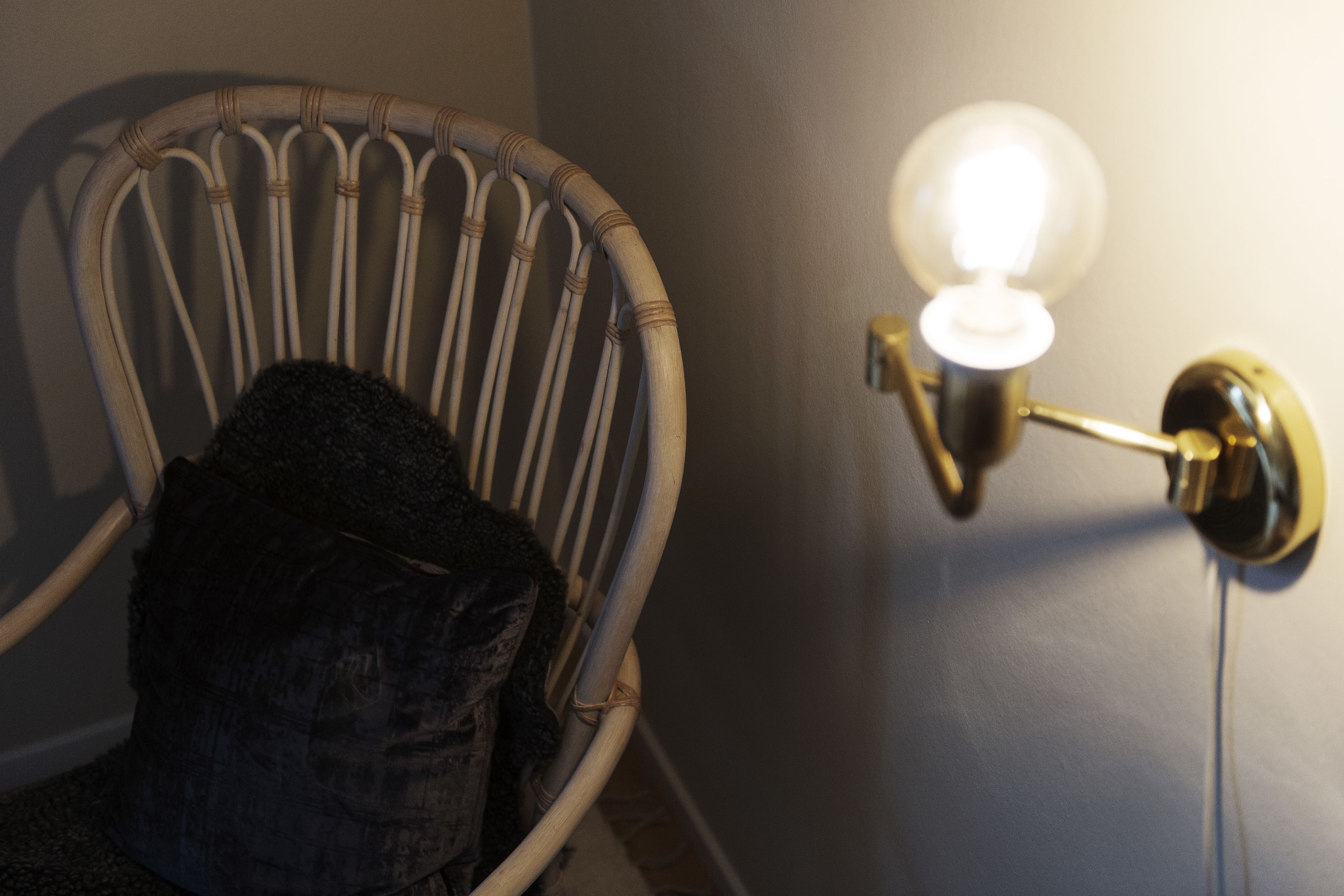A wicker chair with a cushion against a wall, next to a wall-mounted light fixture.