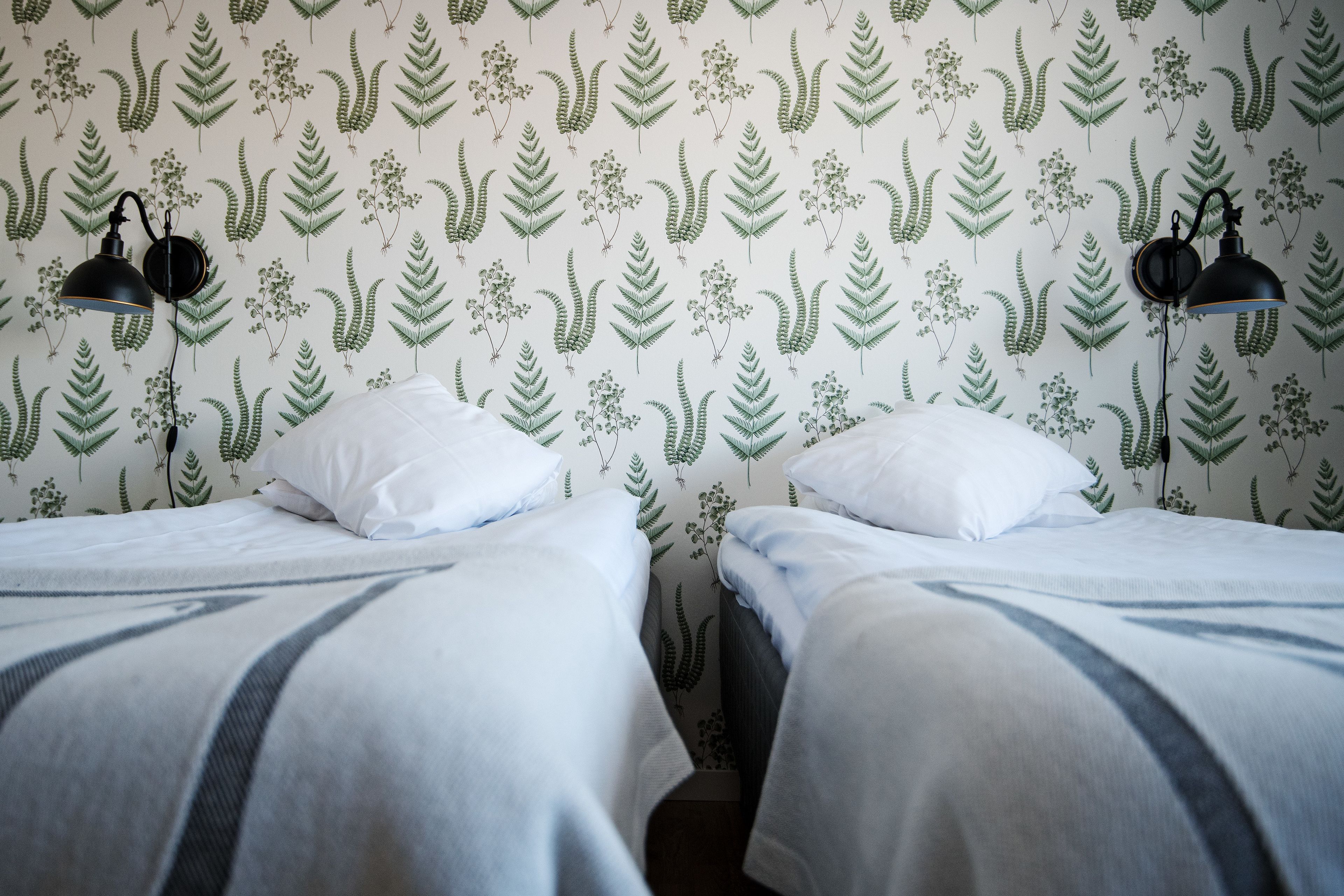 A room with two beds, each with white bedding and a striped blanket. There are two black wall lamps on either side of the beds. The walls are decorated with a green leafy pattern.