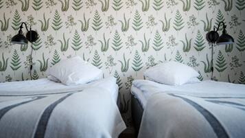 A room with two beds, each with white bedding and a striped blanket. There are two black wall lamps on either side of the beds. The walls are decorated with a green leafy pattern.