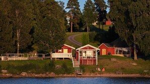 A lakeside cabin with a dock, surrounded by dense forest and a hill in the background. - Två Skyttlar (Orby)