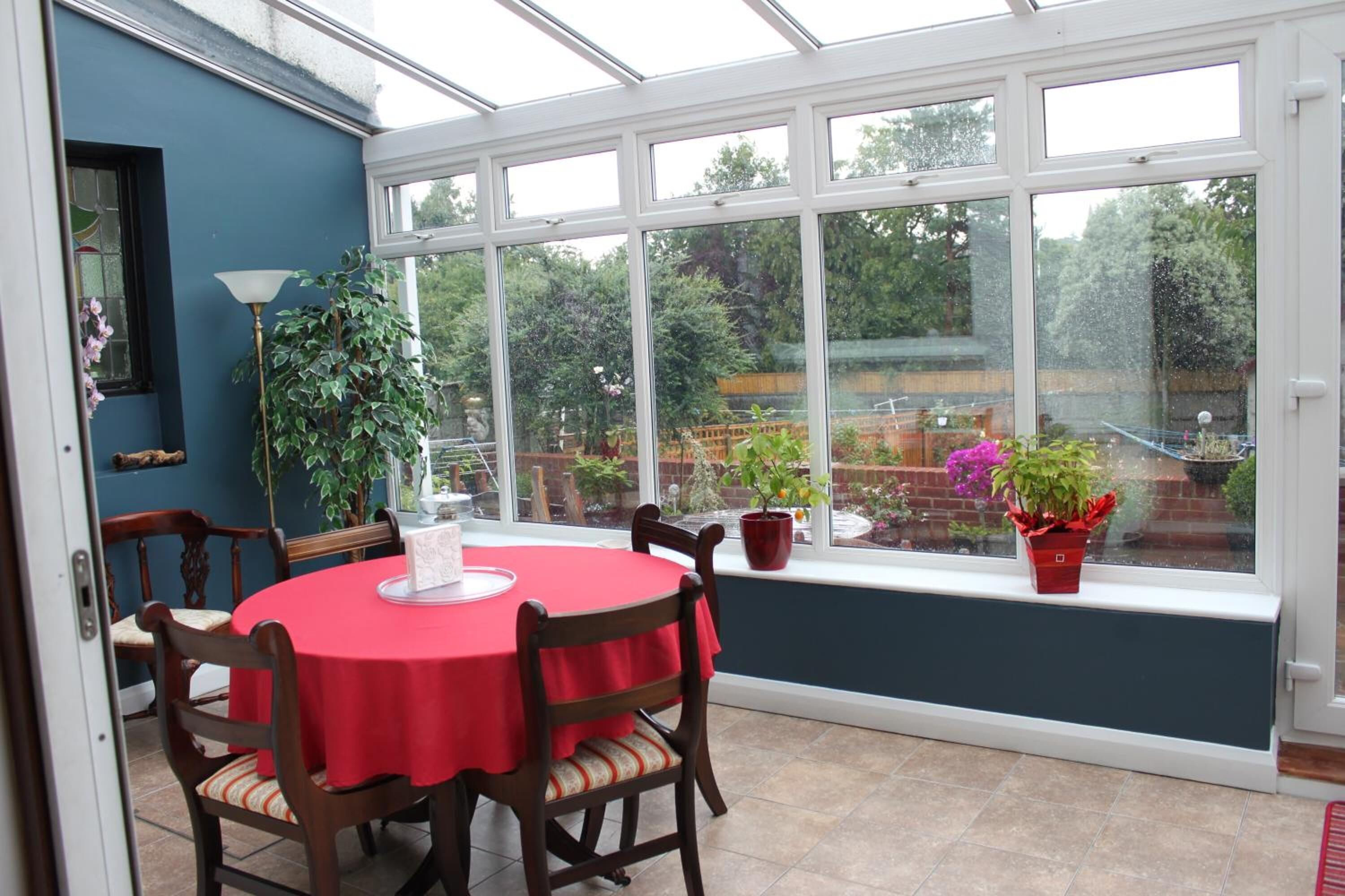 A conservatory with a red table and chairs, large windows, and potted plants.