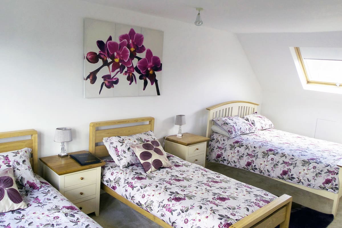Une chambre avec deux lits simples, une table de chevet avec une lampe, une petite table à côté du lit avec un livre, et une fenêtre donnant sur un paysage agréable.