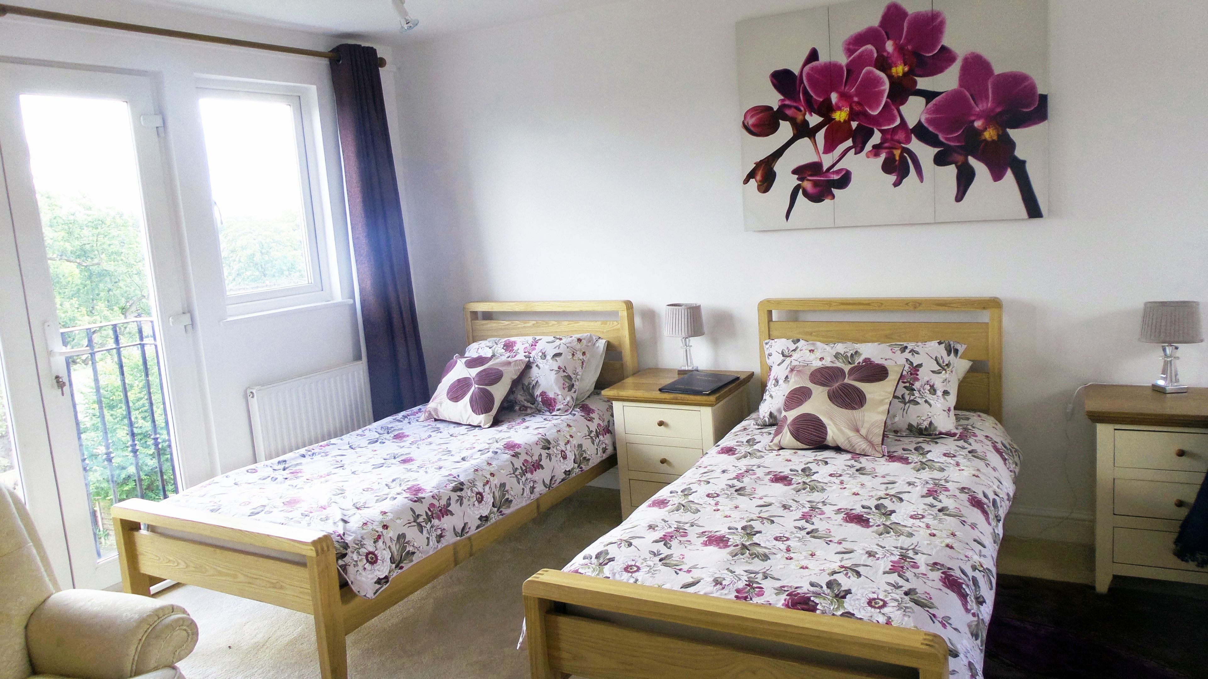 Une chambre à coucher avec deux lits en bois, chacun agrémenté de draps à motifs floraux et d’oreillers assortis. Une table de chevet avec une lampe se trouve entre les deux lits, et une fenêtre avec des rideaux est située sur le côté gauche.