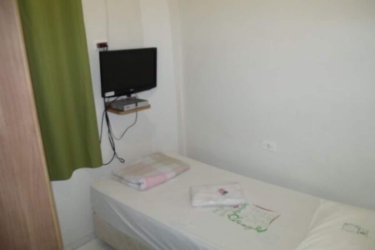 Una stanza piccola con un letto singolo, una televisione su un mobile, uno scaffale e un letto con lenzuolo bianco e cuscino rosa.
