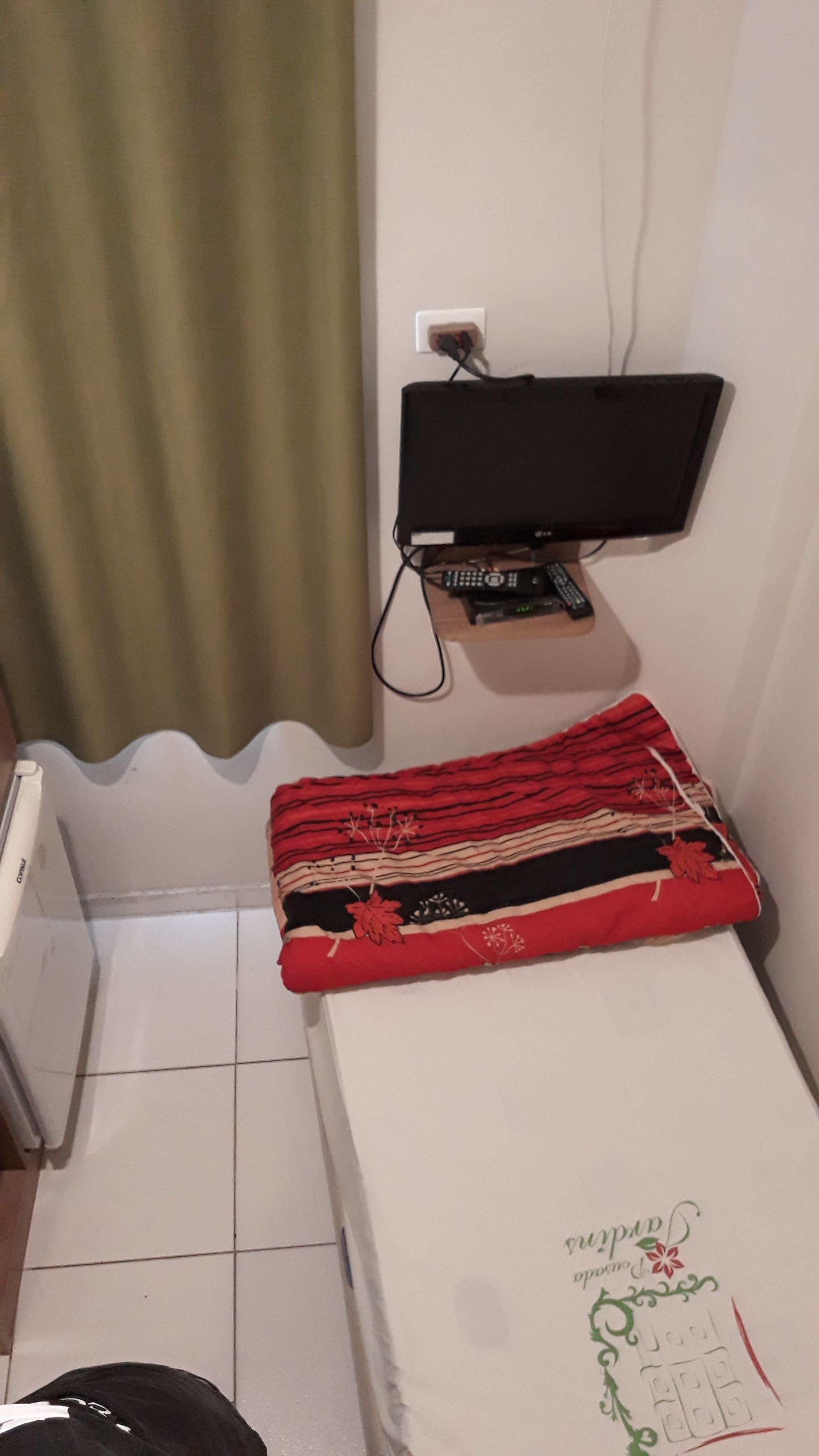 Una habitación pequeña con una cama, un televisor montado en la pared y un enchufe.