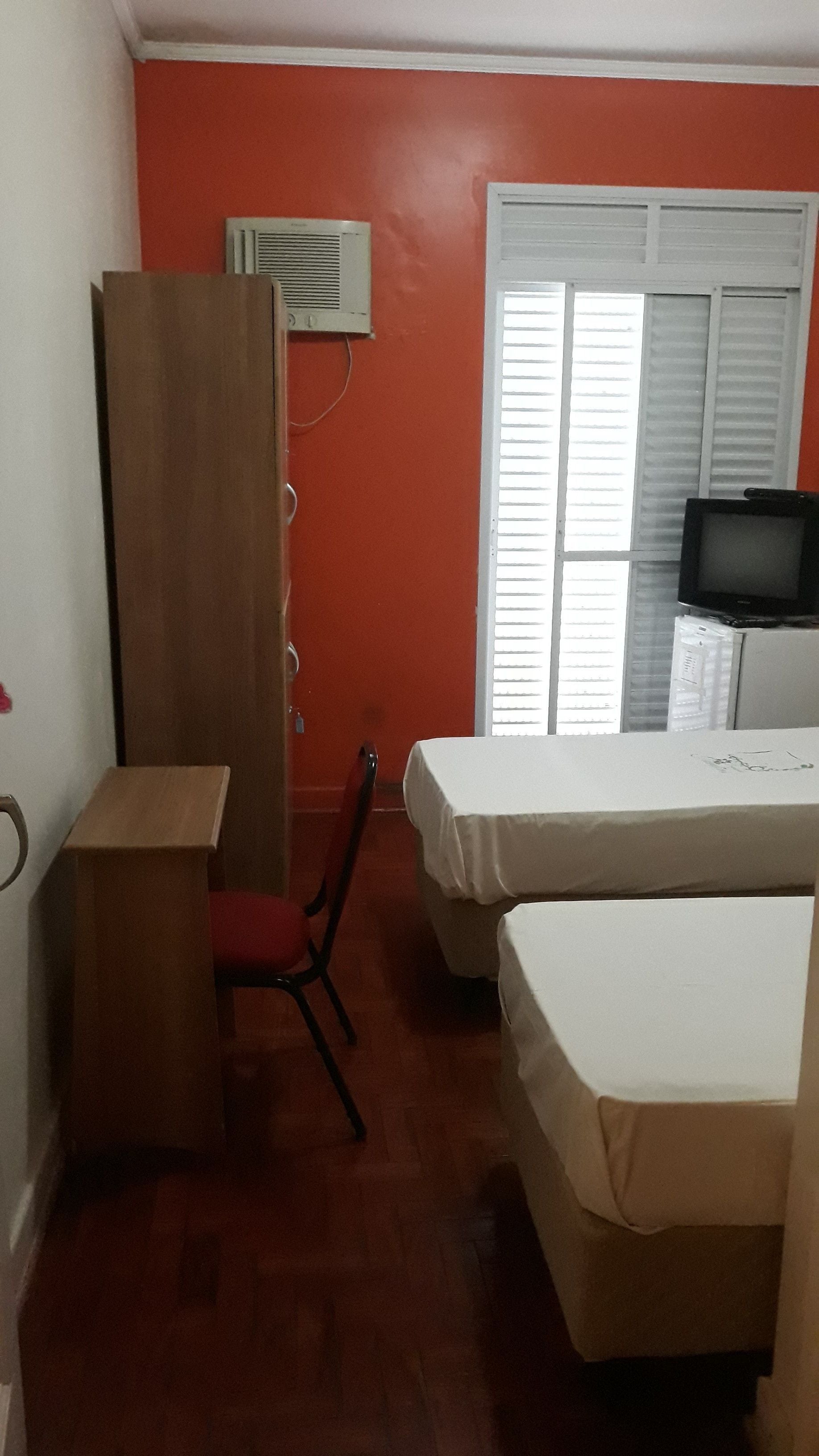 Een kamer met een houten garderobe, een bureau, een stoel, twee bedden met witte lakens, een airconditioning en een televisie op een kast.