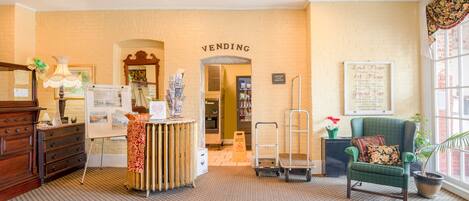 A hotel lobby with a vending machine, a green armchair, a wooden dresser, and a bulletin board.