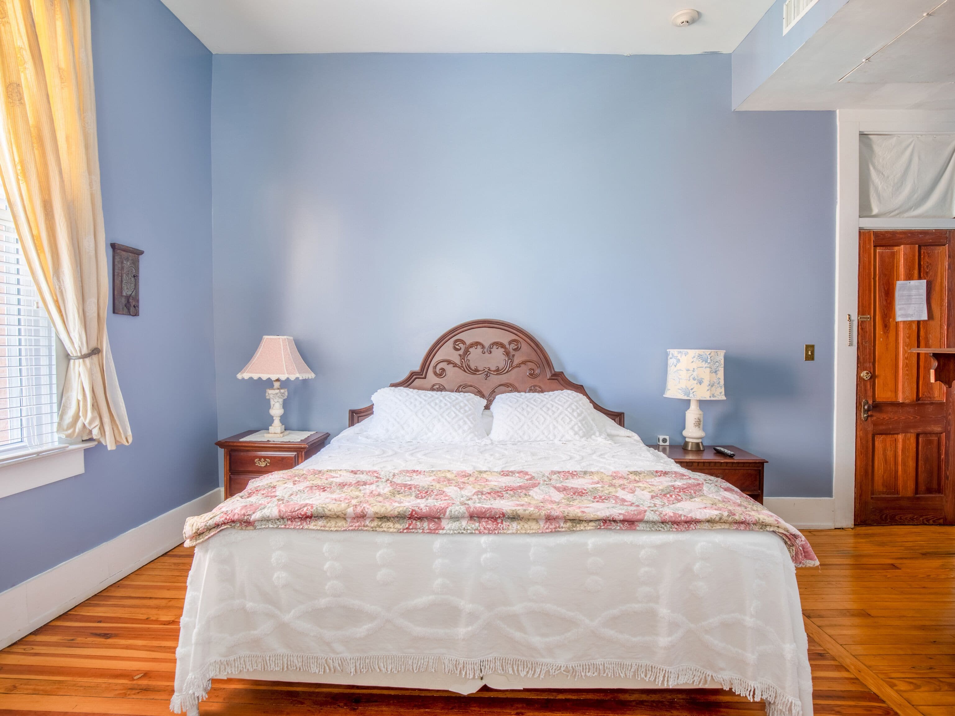 A bedroom with a wooden bed, two bedside tables with lamps, a window with curtains, and a door.
