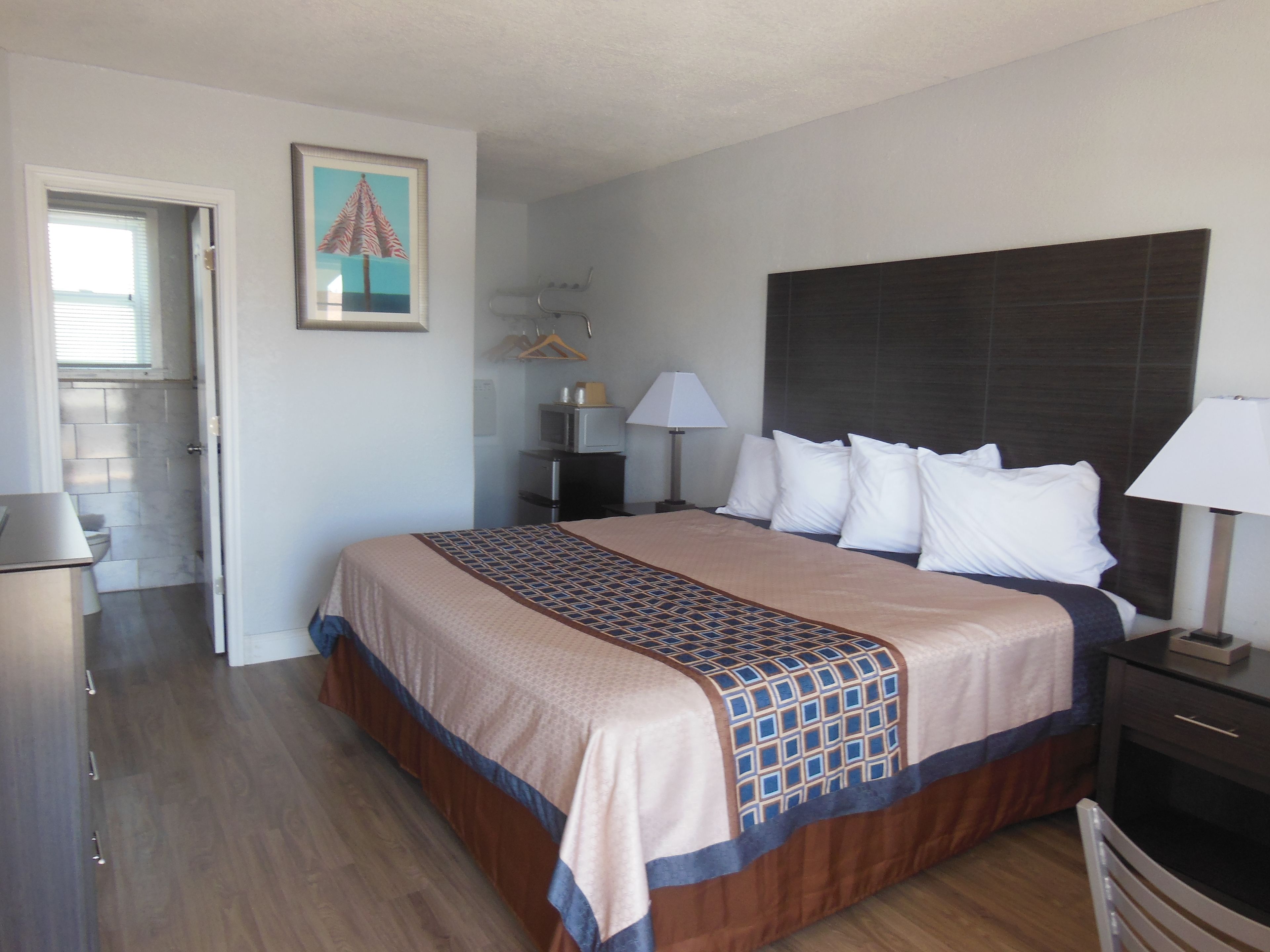 Quarto de hotel com cama, mesas de cabeceira, luminária, quadro emoldurado e vista para o banheiro.