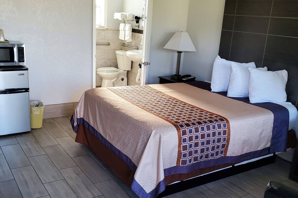 Habitación de hotel con cama, mesita de noche, lámpara, microondas, lavabo e inodoro.