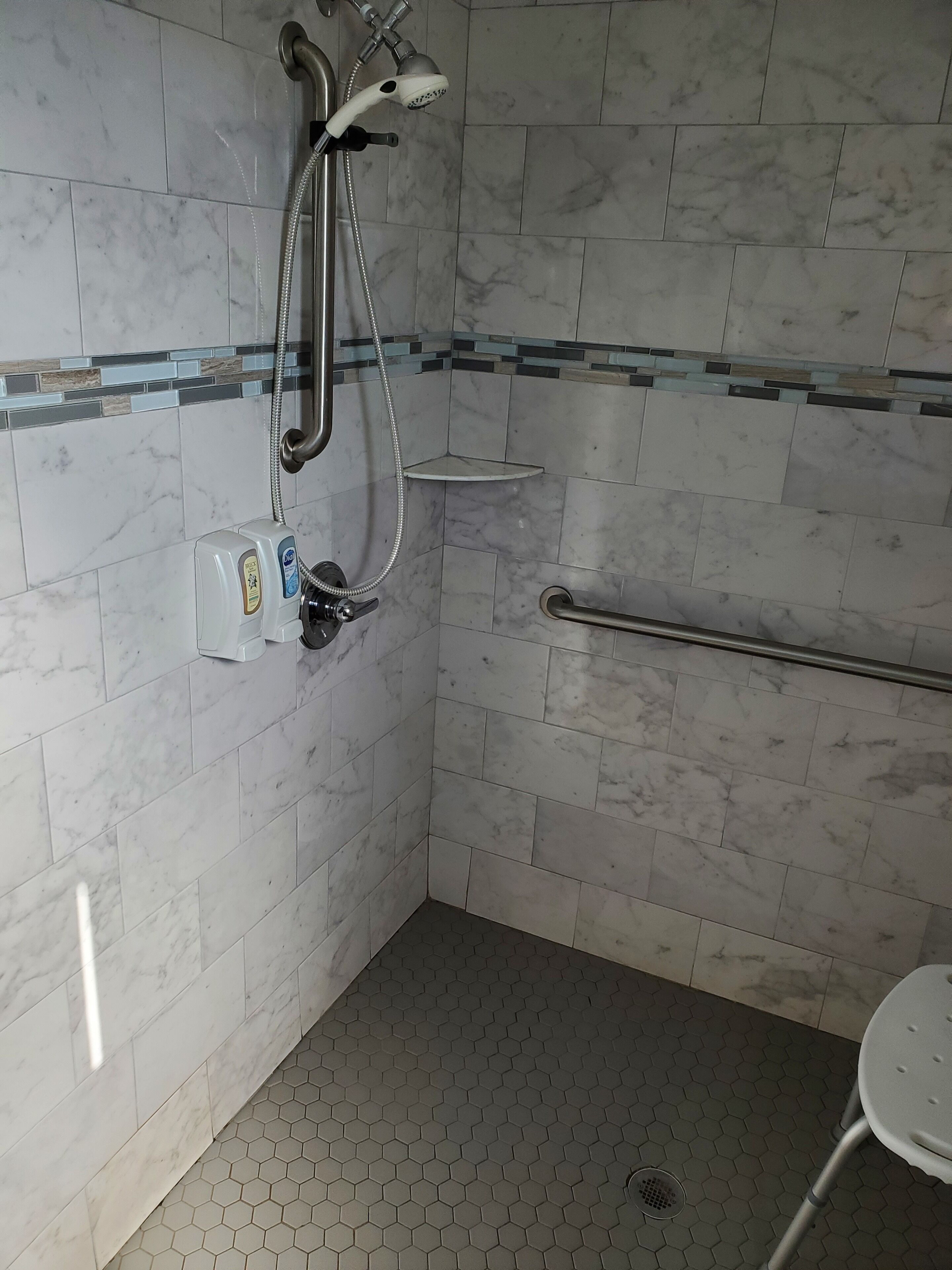 Un baño con baldosas de mármol blanco, una ducha con cabezal de ducha manual, un asidero y un estante pequeño.