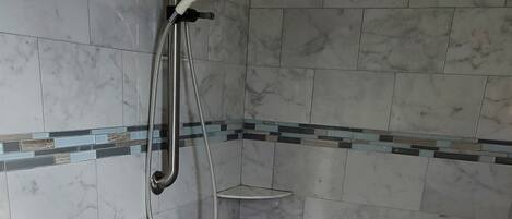 Un baño con baldosas de mármol blanco, una ducha con cabezal de ducha manual, un asidero y un estante pequeño.
