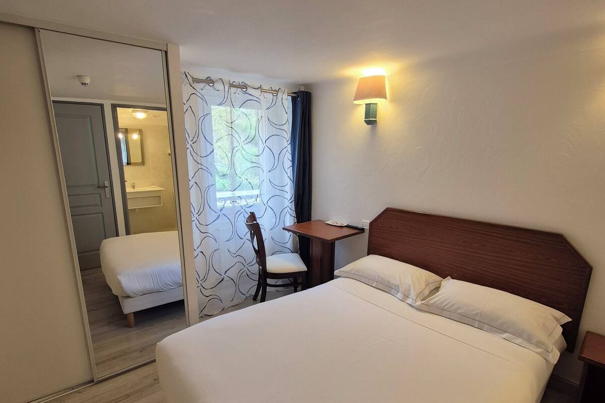 Ein Hotelzimmer mit Bett, Schreibtisch, Stuhl, Spiegel, Fenster mit Vorhängen und einer Wandlampe.