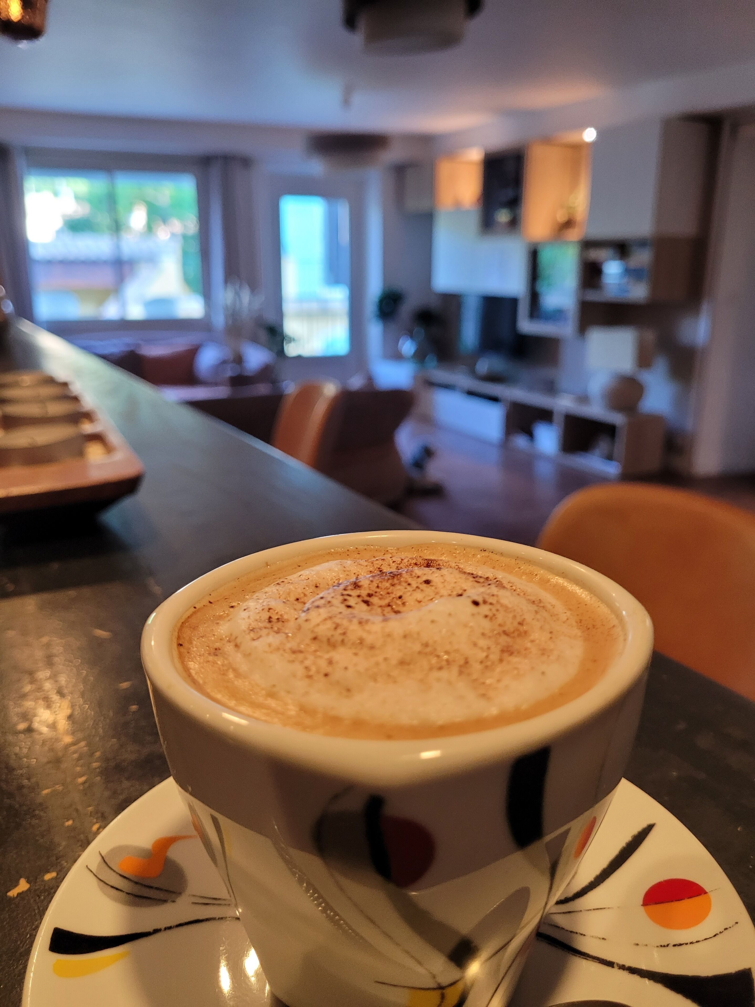 Una tassa de cafè sobre un plat, una cullera, una taula i vista d'un interior modern amb seients i zona de cuina.