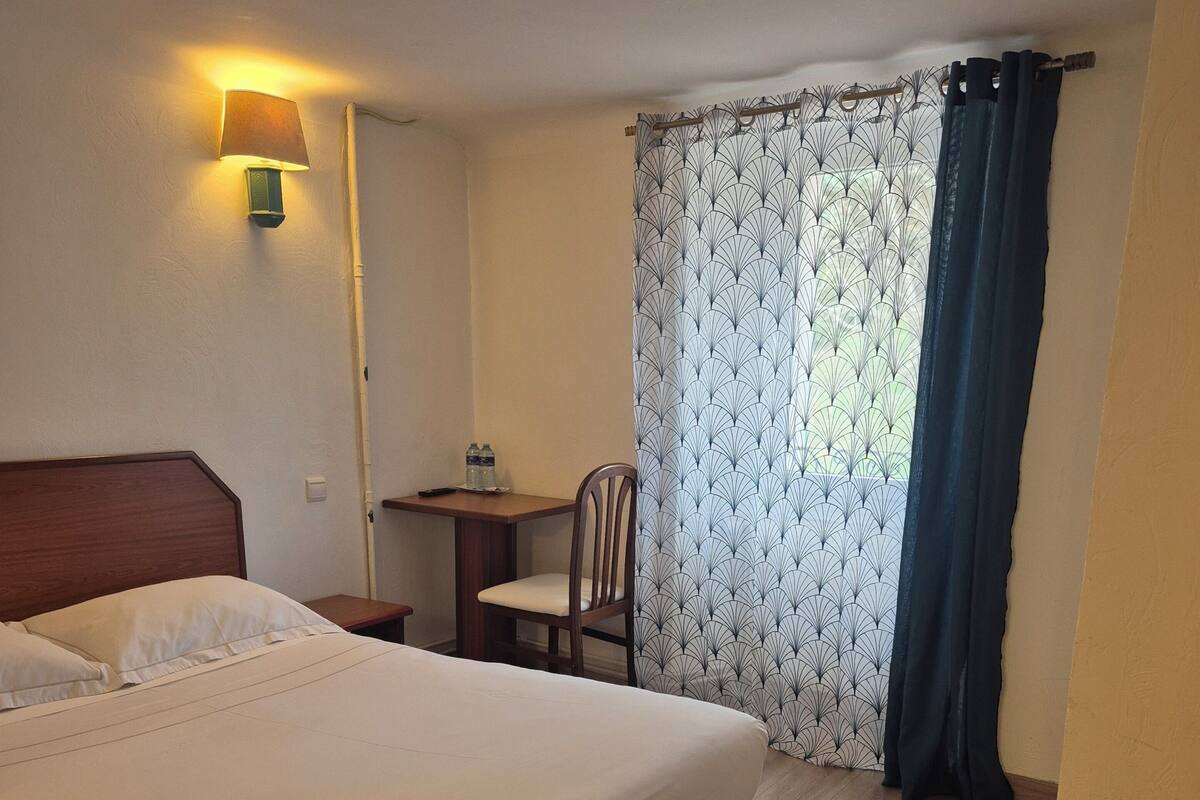 Ein Hotelzimmer mit einem Bett, einem Schreibtisch, einem Stuhl, einer Lampe und einem Vorhangfenster.