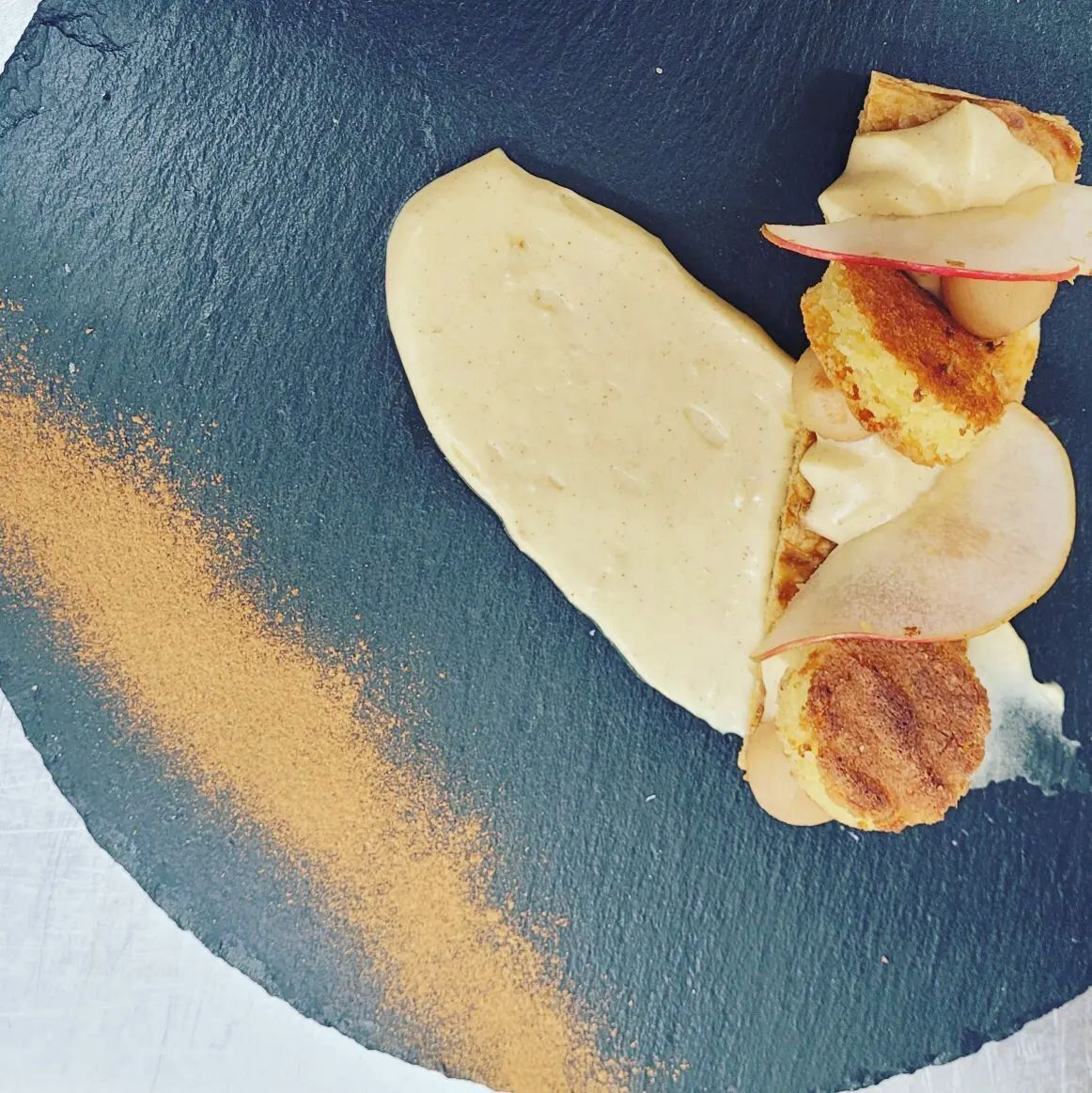 Un plato con un postre suave y pálido, rodajas de manzana y una porción de pastel sobre una superficie de pizarra oscura.