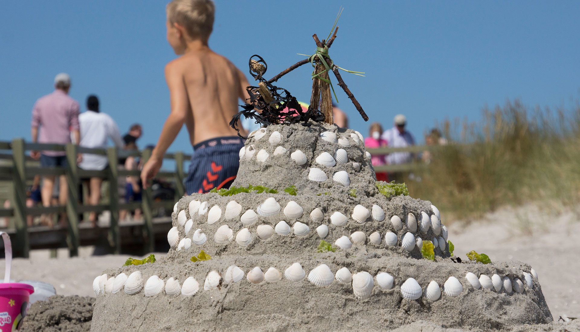 Et sandslot med søskaller og en figur øverst, beliggende på en strand med folk i baggrunden.