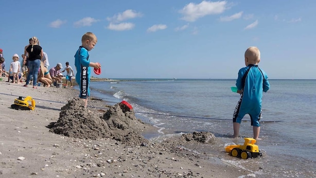 Escena de playa con niños jugando cerca del agua, una duna de arena y vehículos de juguete.