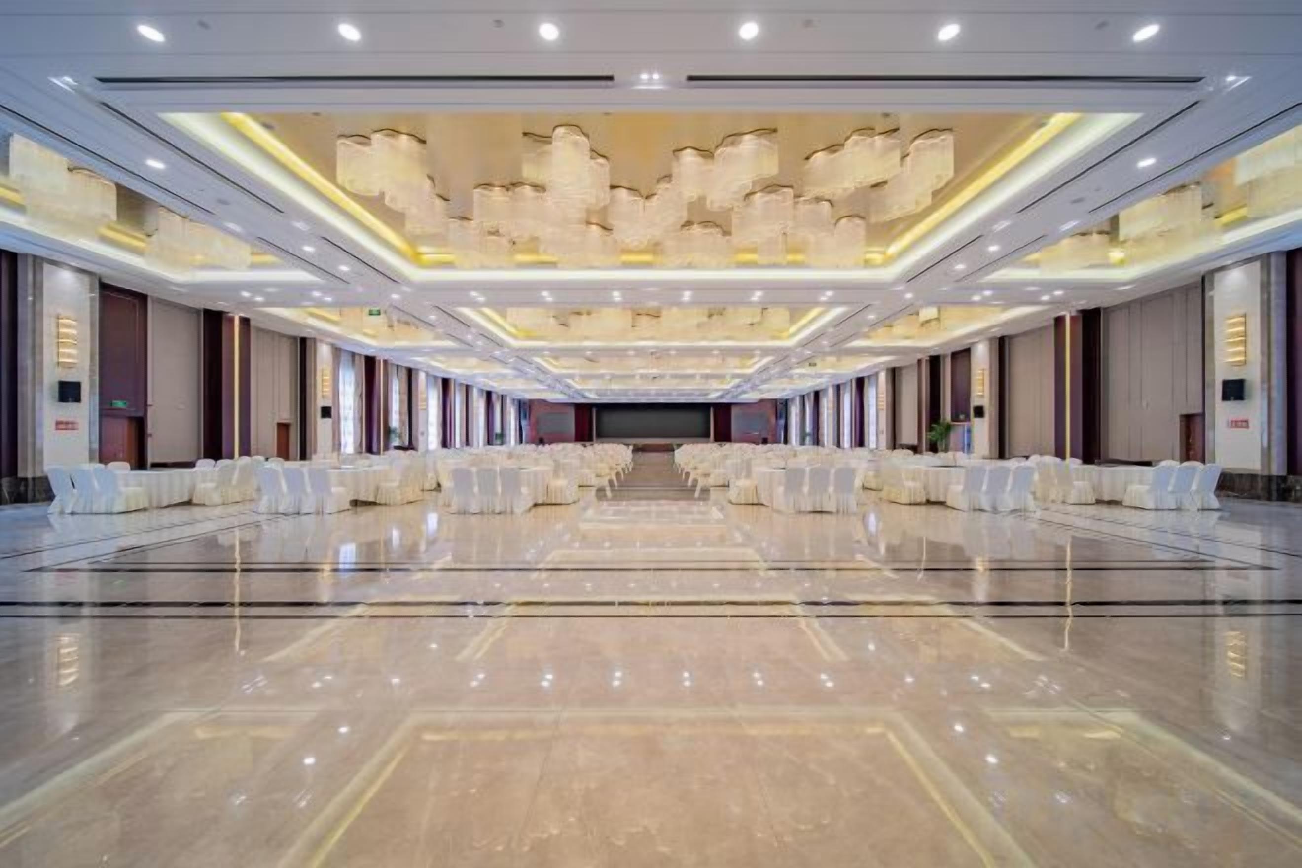 A spacious banquet hall with rows of white round tables and chairs, a polished marble floor, and a high ceiling with recessed lighting.