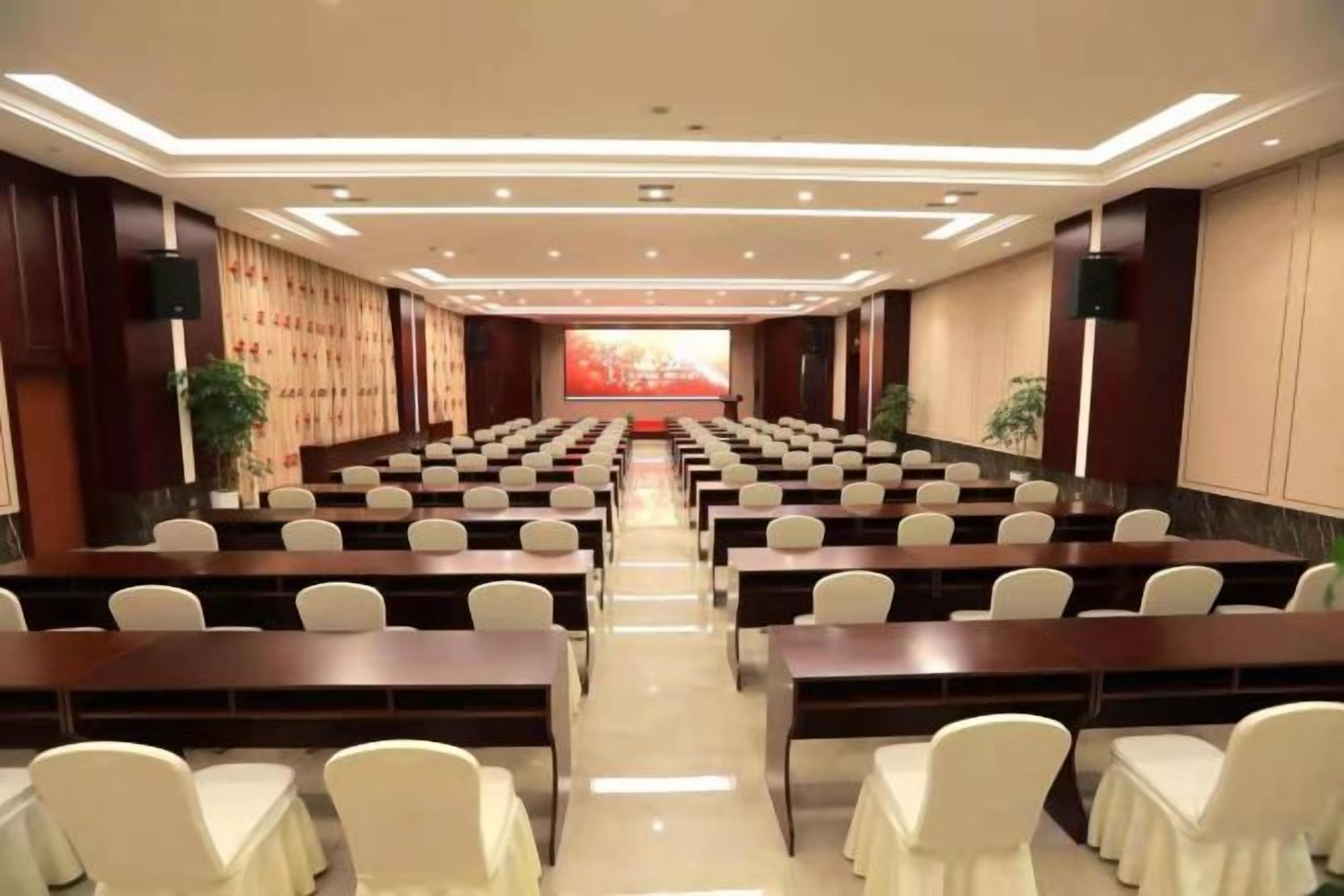 Un salón de conferencias con filas de mesas y sillas, un escenario al frente y una gran pantalla.