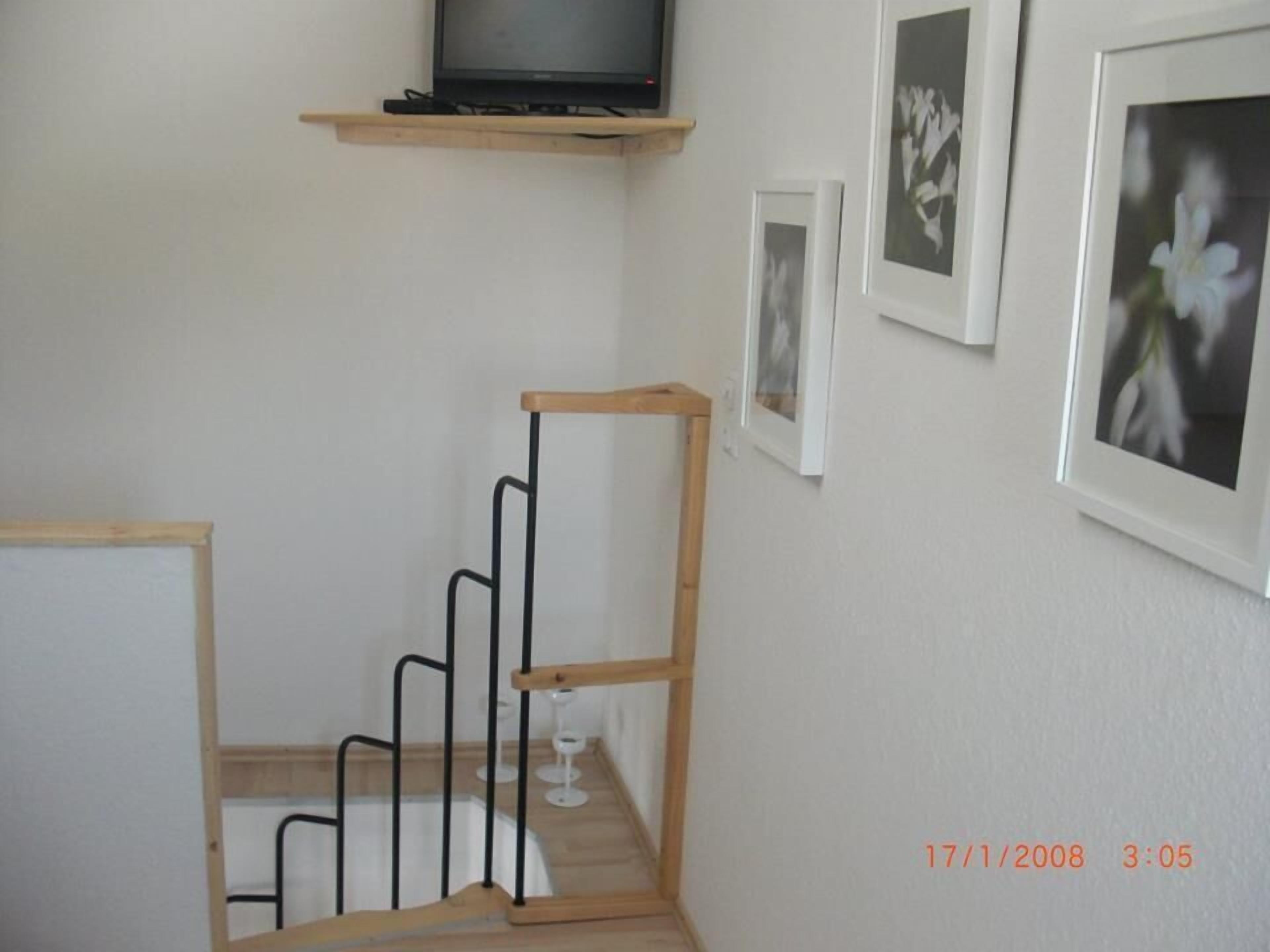 Una escalera con barandilla de madera y balaustradas de metal negro, un televisor en un estante encima y cuadros enmarcados en la pared.