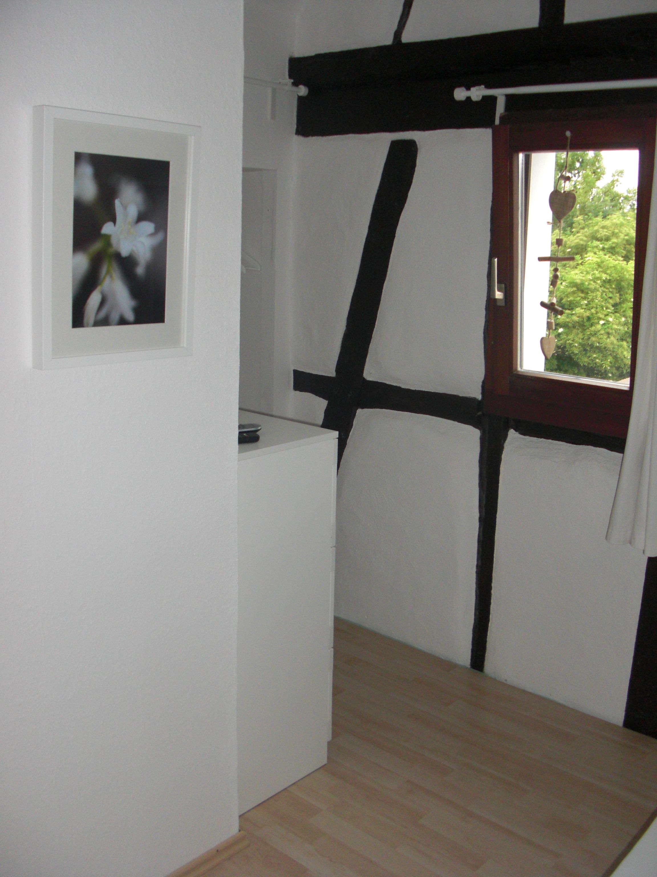 Una habitación con piso de madera, pared blanca con un cuadro enmarcado y ventana con vista a vegetación.