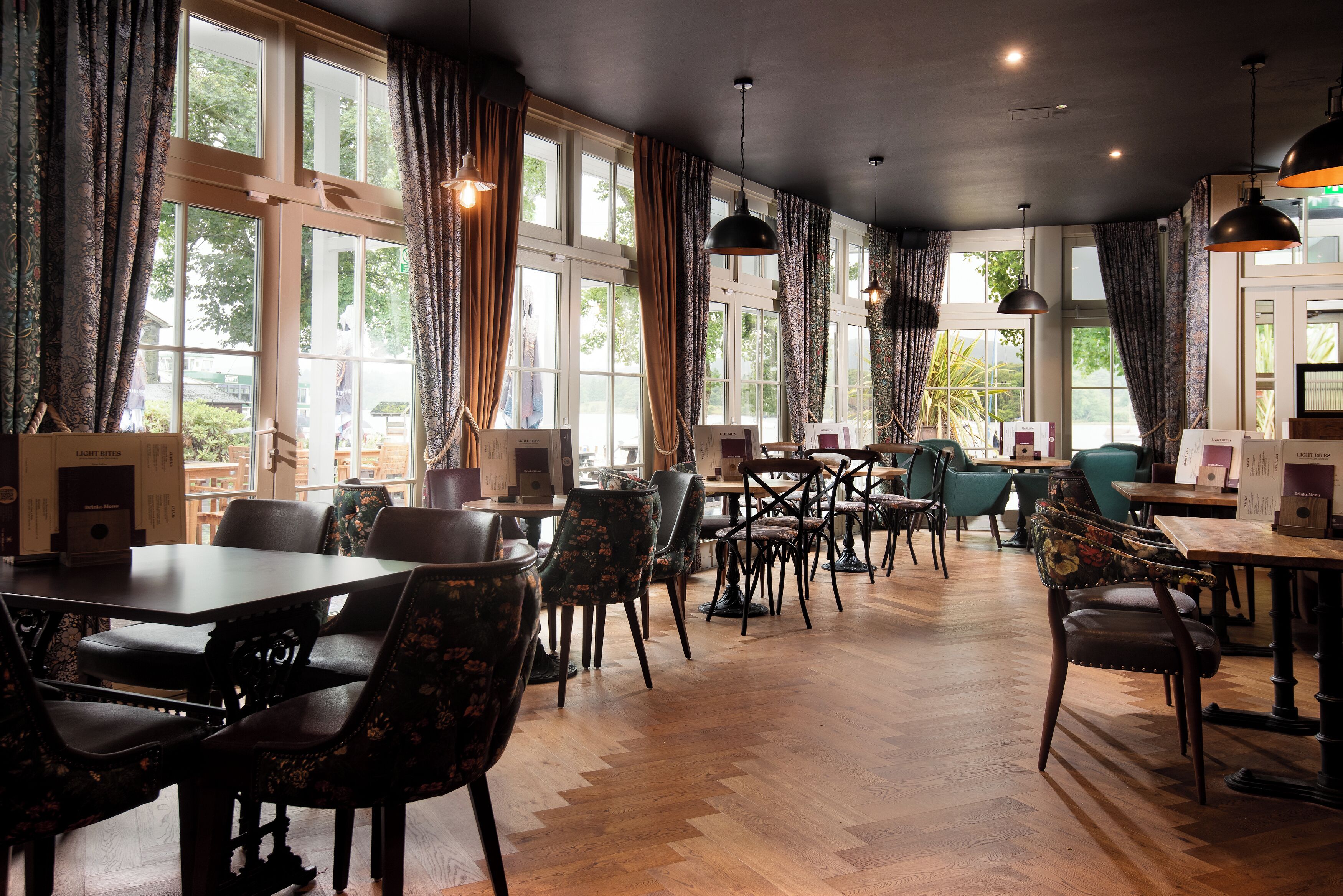 A spacious dining area with multiple tables and chairs, large windows with curtains, and hanging pendant lights.