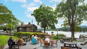 An outdoor seating area with wooden tables and benches, overlooking a body of water and a stone building. - The Waterhead Inn - The Inn Collection group (Ambleside)
