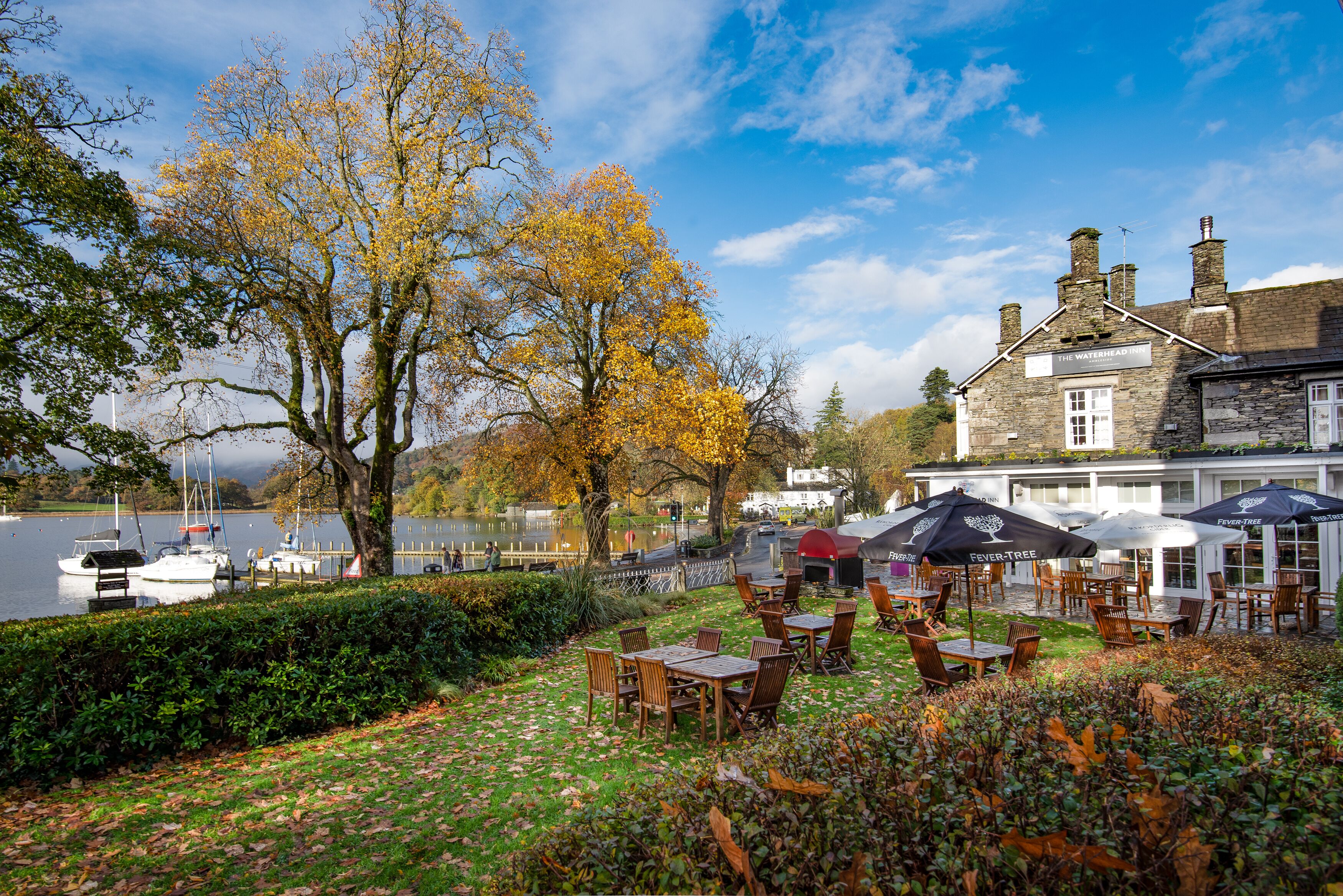 A riverside outdoor seating area with tables and chairs, a stone building labeled "The Waterhead Inn," and boats docked in the background.