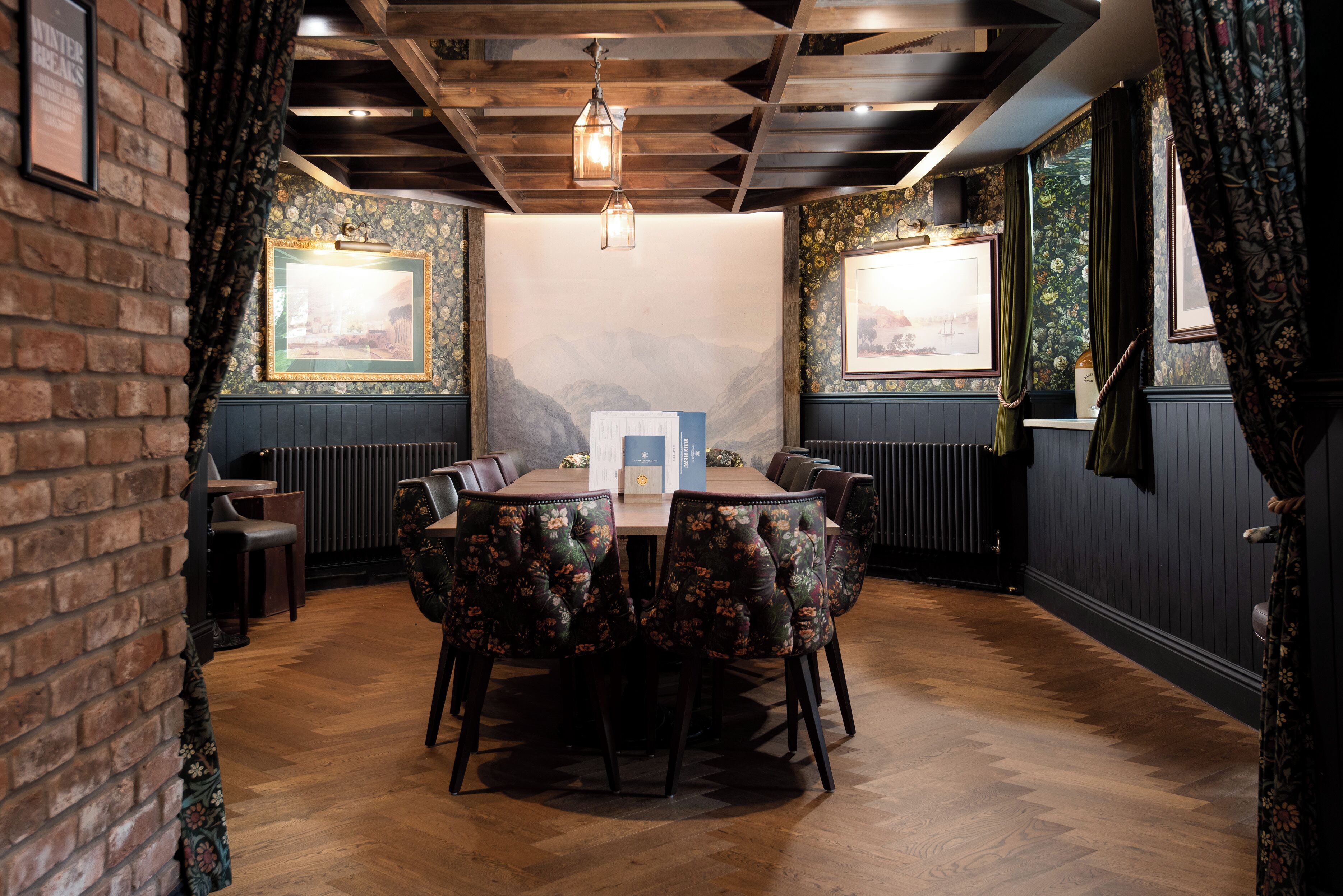 A dining area with a wooden table and floral upholstered chairs, brick walls, and framed pictures on the walls.